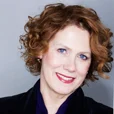 Liz Ann Macgregor has short, curly red hair. Her head is tilted to the left and a black button down shirt. She has fair skin, blue eyes and is smiling.