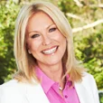 Kerri-Anne Kennerley is smiling with her head tilted to the right. She has straight, blonde hair and wears a white jacket over a pink button down shirt.