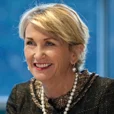 Wendy Machin has short blonde hair with lighter highlights. She is smiling and wears pearl drop earrings, a pearl necklace and a black, lace top.