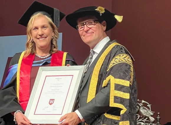 Caroline Hutchinson accepted her Honorary Award from Western Sydney University. She and a man stand on stage in robes holding the framed certificate.