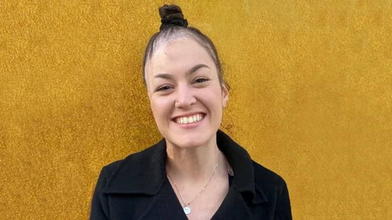 Sophie Delezio is smiling . Her dark hair is pulled back and she is wearing a black shirt. The background is a mustard yellow.