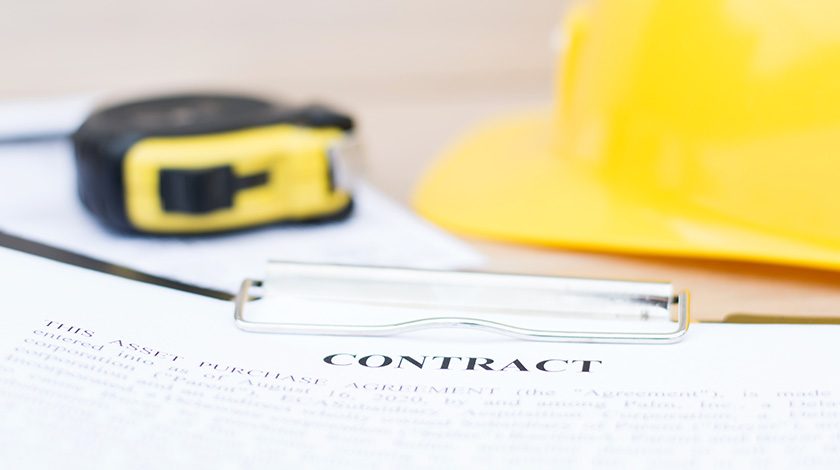 A contract, tape measure and yellow hard hat lay together on a flat surface