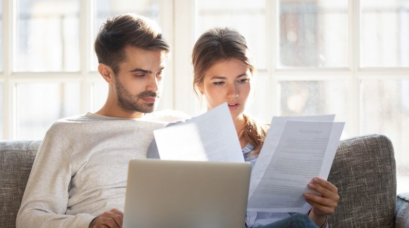 Millennial married couple sitting on couch with computer and documents, reading received formal letter from bank loan mortgage information, contract terms and conditions discuss papers working concept