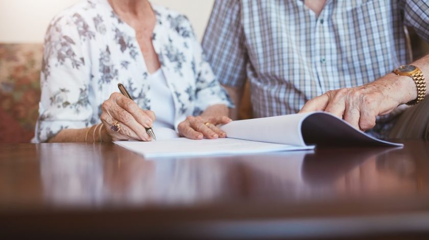 Close up shot of senior woman signing documents with her husband. Elderly caucasian man and woman sitting at home and signing some paperwork, focus on hands.