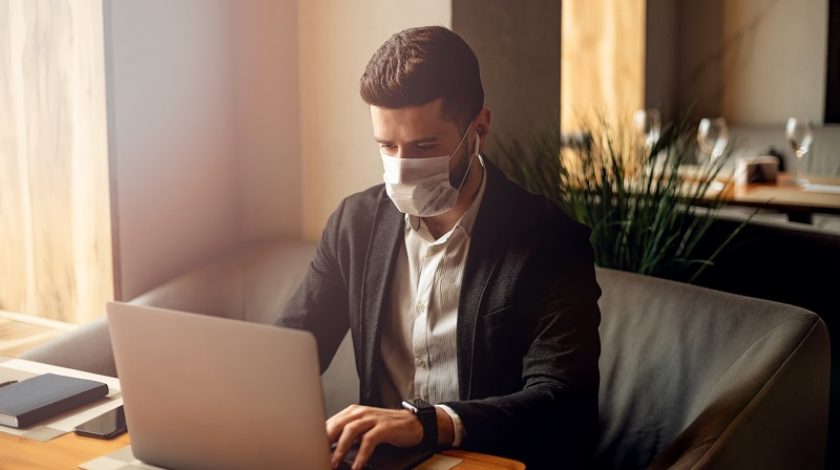 Caucasian adult bearded man indoors in cafe. Lifestyle concept photo with copy space. Picture with handsome guy who using protective face mask. Portrait with gray laptop