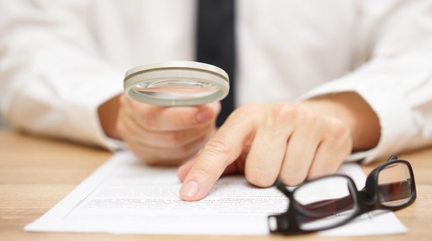 A man's hand holds a magnifying glass over a contract on a table