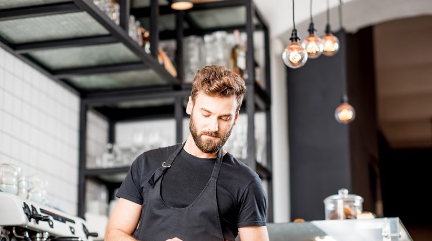 Handsome waiter in black t-shirt working with tablet standing at the bar of the modern cafe interior